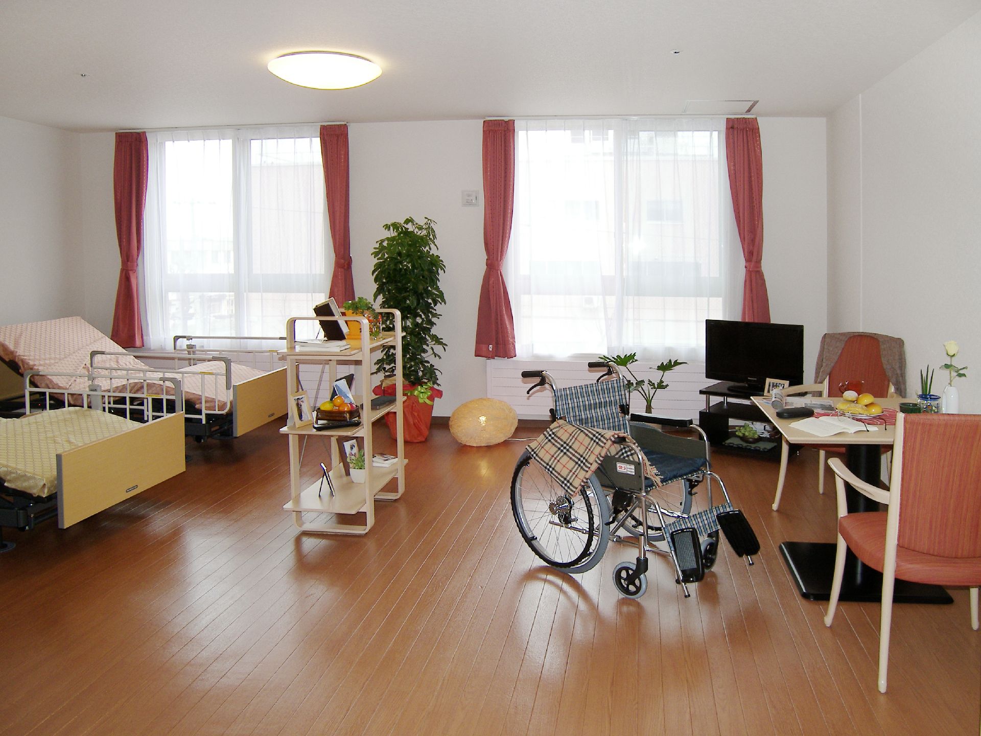 右側にはダイニングテーブルセットやテレビ、車いすがあり、左側には寝具がセットされたベッドが2台あり、観葉植物が飾られている。