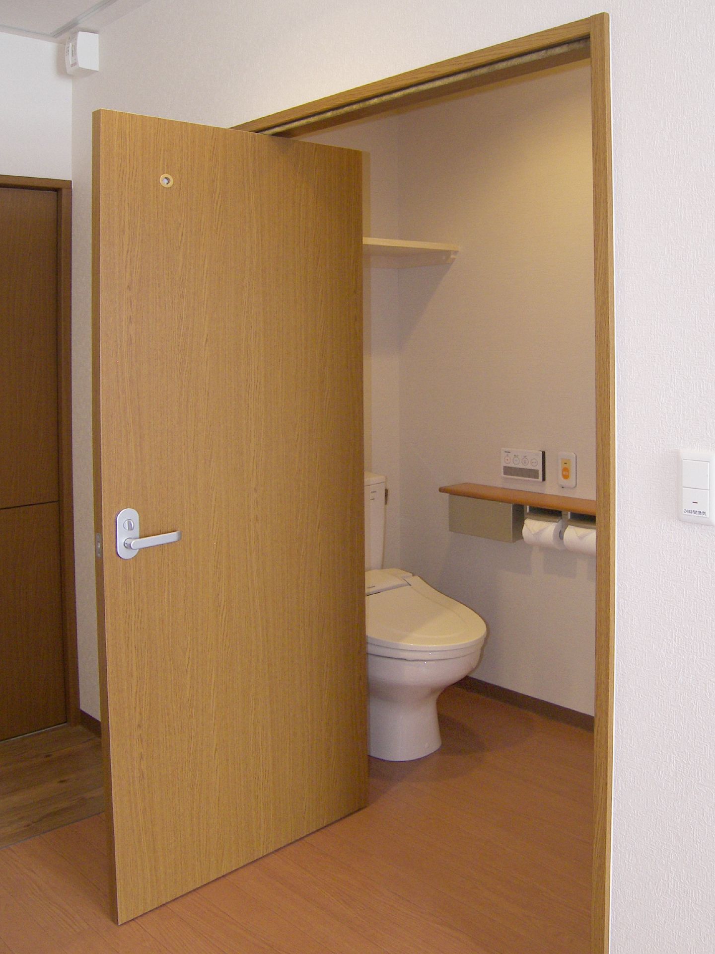 大きなドアが付いたトイレには収納棚があり、壁にはオレンジの呼び出しボタンや2連のトイレットペーパーホルダーが設置されている。