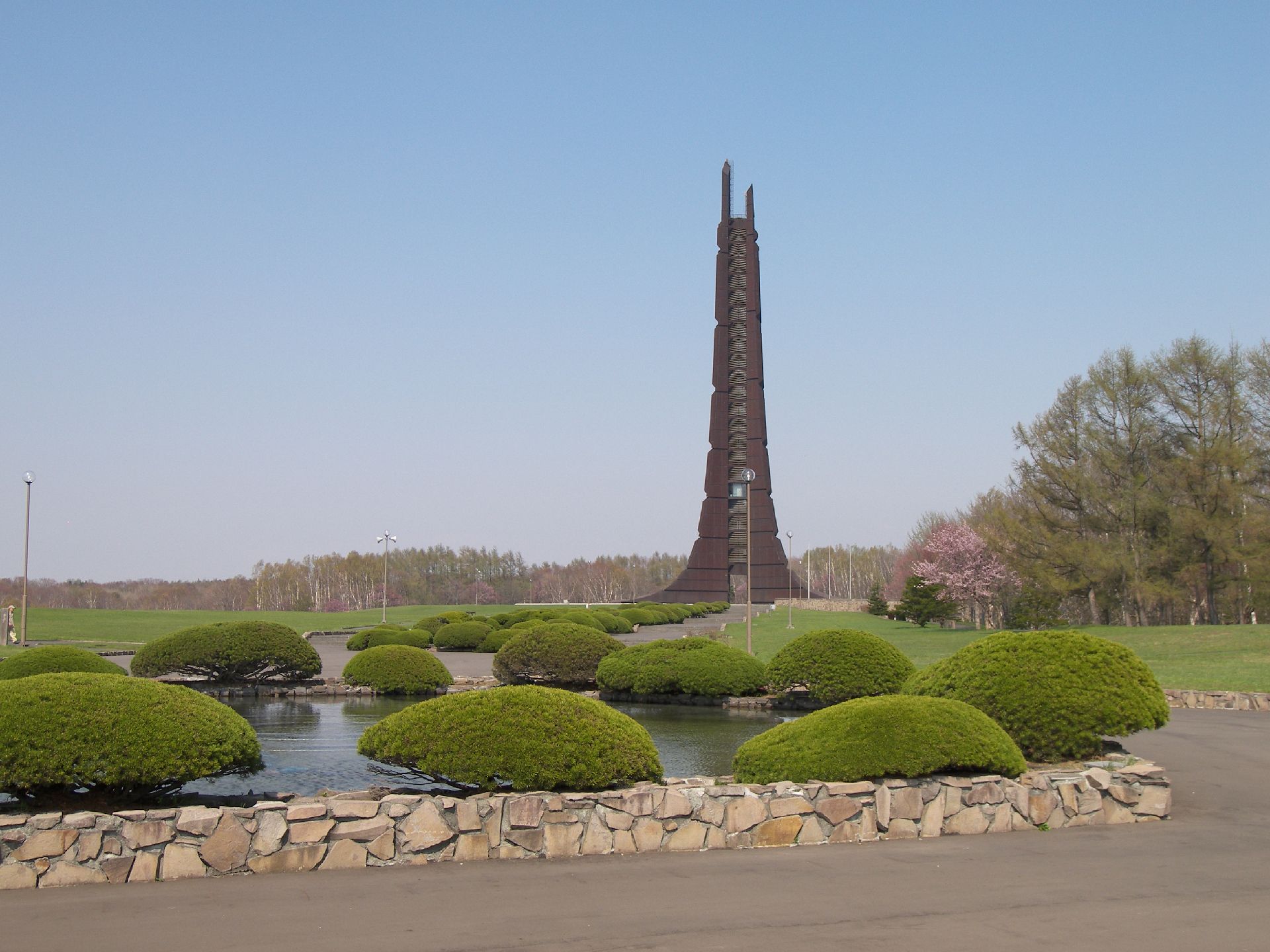 池があり、芝生が敷かれた公園の一角に立てられたダークブラウンの塔。周辺には木もたくさん植えられている。