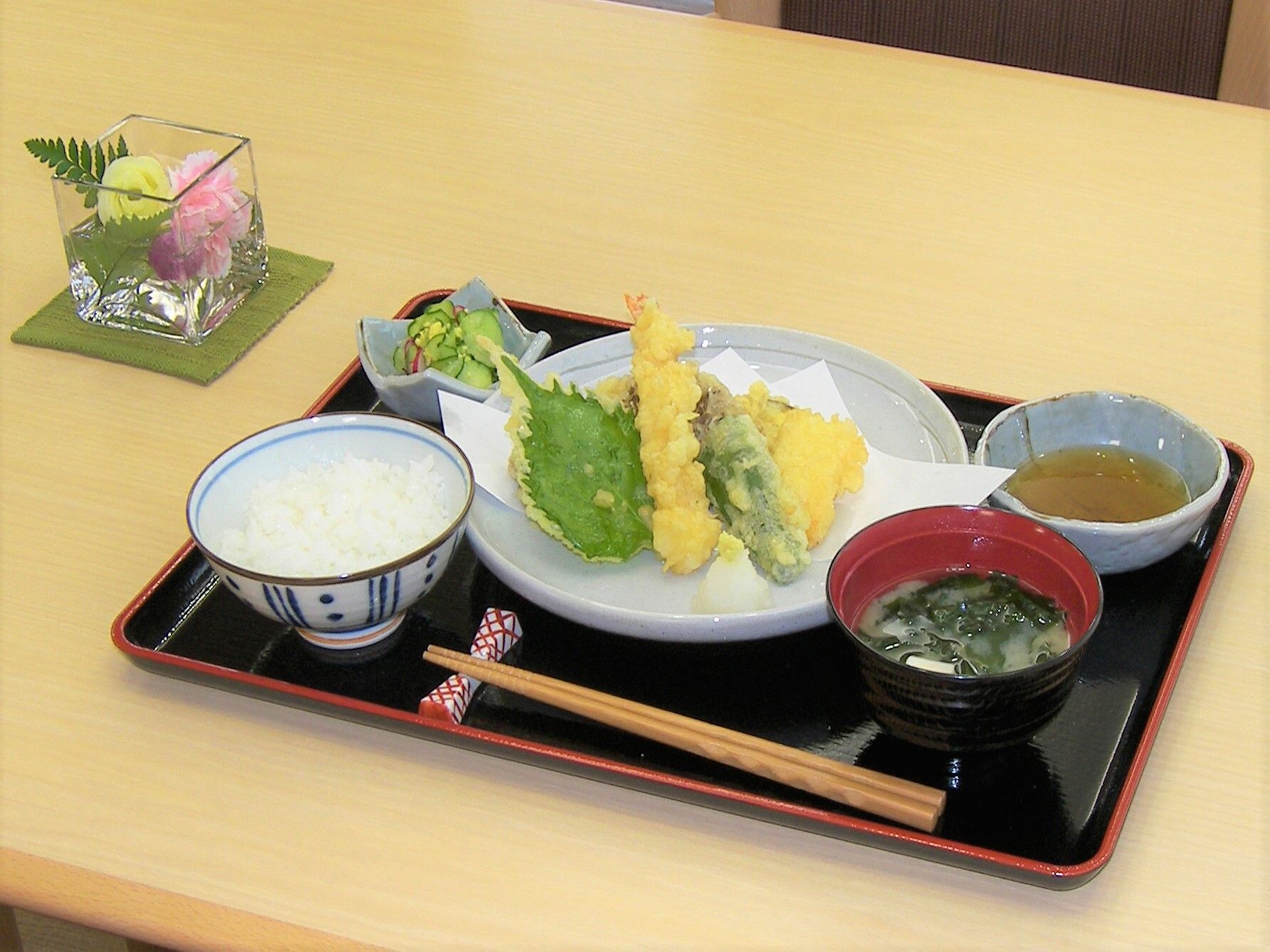 天ぷらの盛り合わせやみそ汁、白いご飯、野菜の和え物などが並んだ黒いトレーには、箸と箸置きも置かれている。