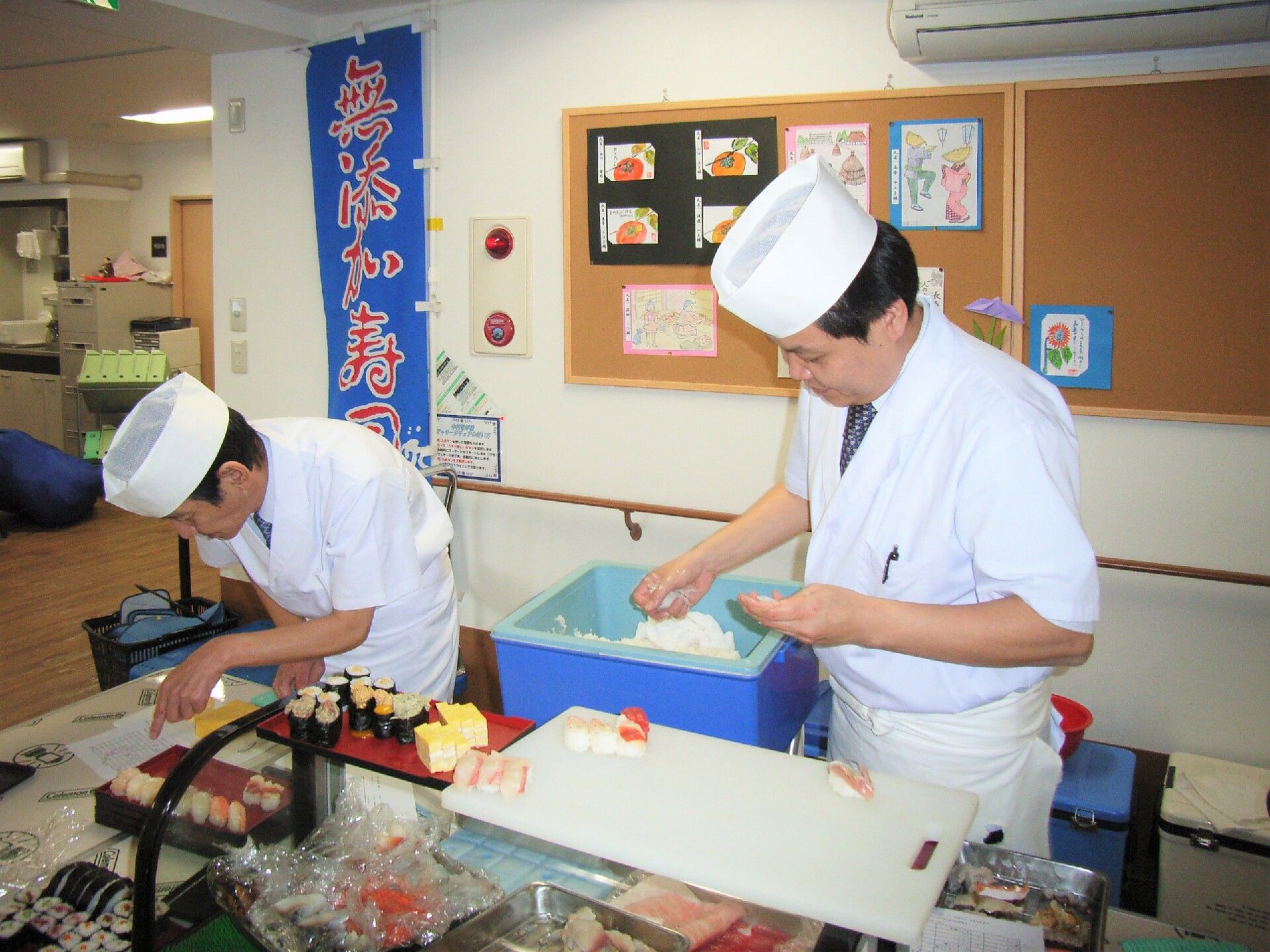 「無添加寿司」と書かれたのぼりが立ち、２名の寿司職人が寿司を握っている様子。卵やエビ、鉄火巻きなどが並んでいる。