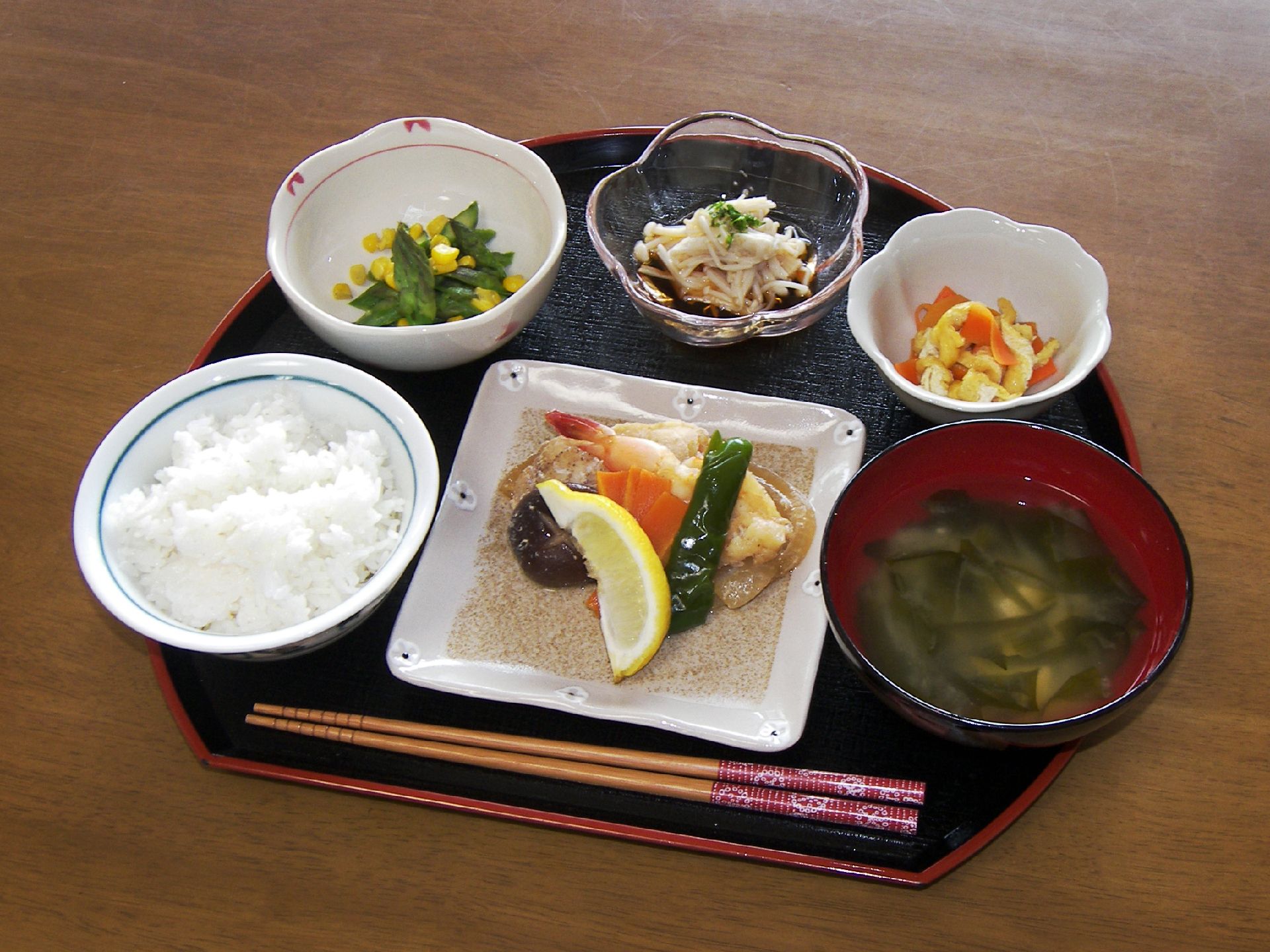 レモンが添えられた野菜と魚介類のグリルのようなものに、白いご飯とみそ汁、3種類の野菜の小鉢が並んだ黒い盆。