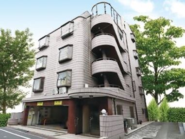５階建てのグレーの建物。屋外階段が付き、建物の周辺には大小の木が植えられている。