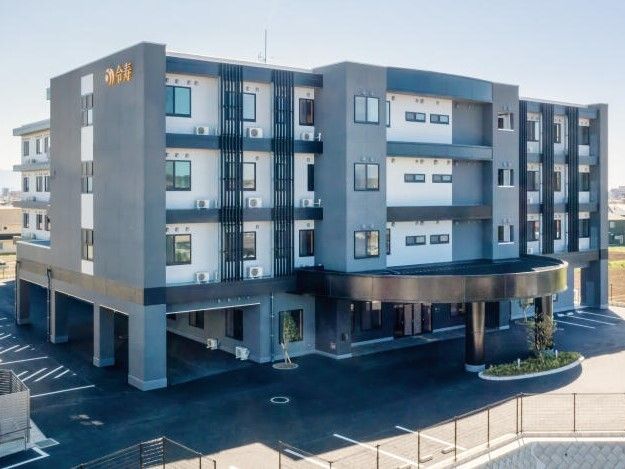 ４階建ての建物。グレーをメインとしていて、外壁にはホーム名が書かれ、車寄せが玄関につ、坂を下りると駐車場が広がっている。