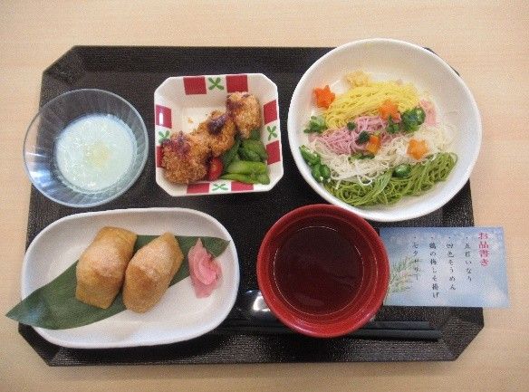 白いご飯に味噌汁、焼き酒や冷ややっこ、ヒジキの煮物、梅干しなどが盛られた器が並んだ木製のテーブル。箸置きには箸が置かれている。