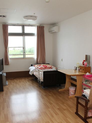 カーテン付きの大きな窓が付いた明るいフローリングの部屋には、寝具付きのベッドや木製のテーブルなどがある。