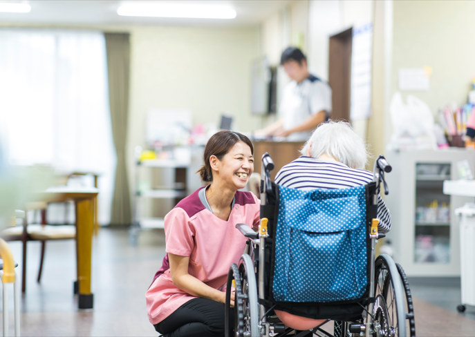 白いゆったりとした服を着た高齢女性が車いすの高齢男性に寄り添い、笑顔で話している。