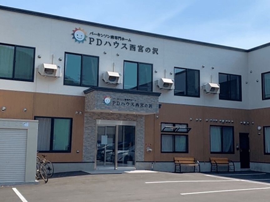 ２階建ての建物。1階部分はベージュで、2階部分は白。玄関には屋根が付いていて、ベンチも置かれている。