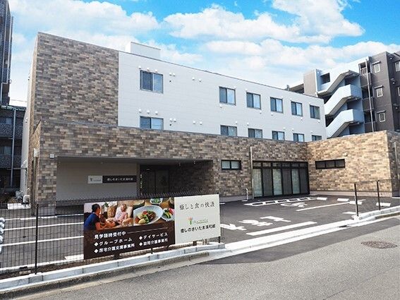 白とブラウン系の3階建ての建物は横に長く、大きな駐車場も付き、建物周辺には黒いフェンスがある。