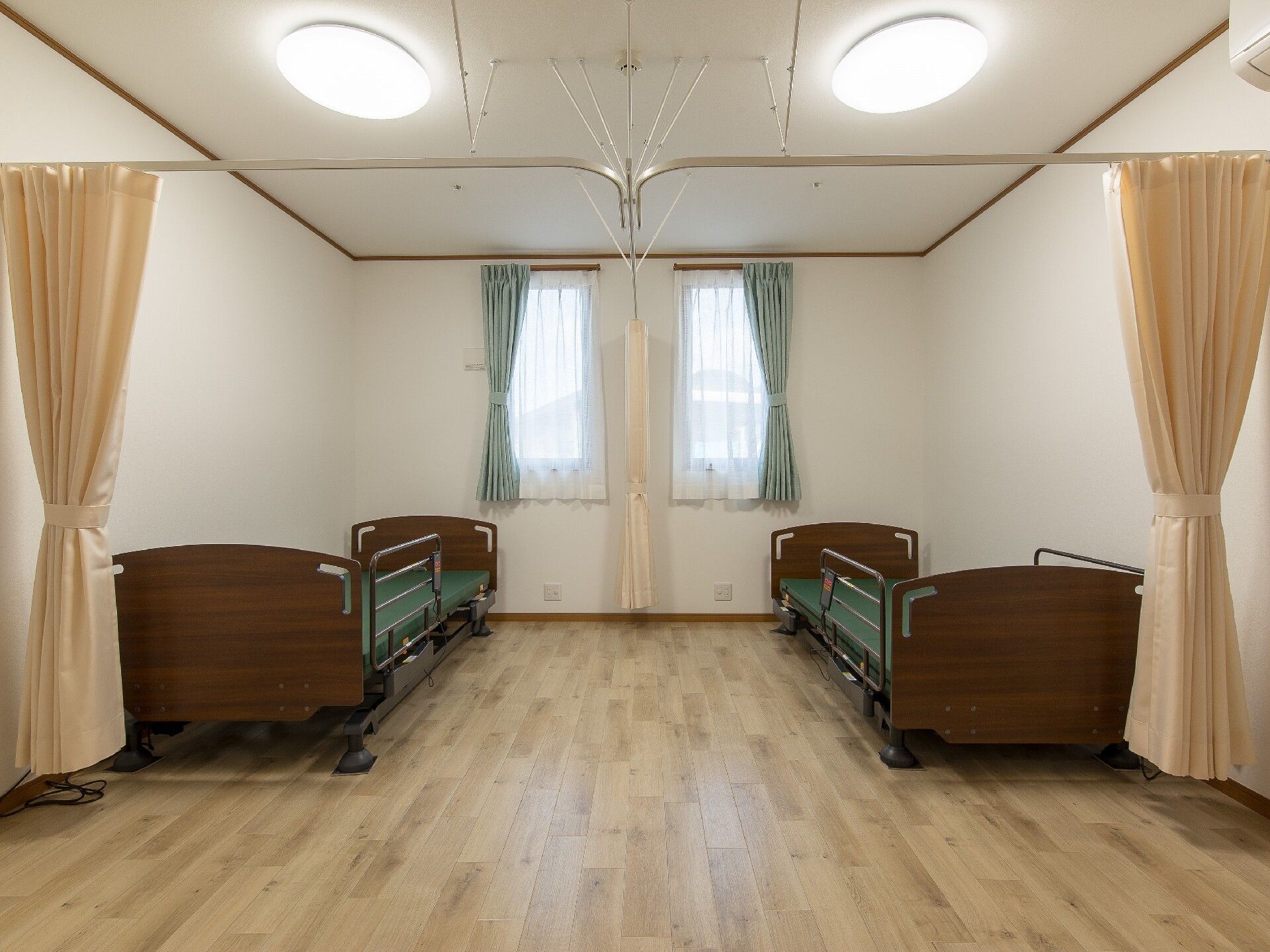 フローリングの部屋には介護ベッド２台やカーテンが付いた窓、エアコンがあり、中央にカーテンが設置されている。