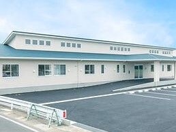 白い外壁でグリーンの屋根が付いた平屋建ての建物。敷地内には広い駐車場がある。車は止まっていない。