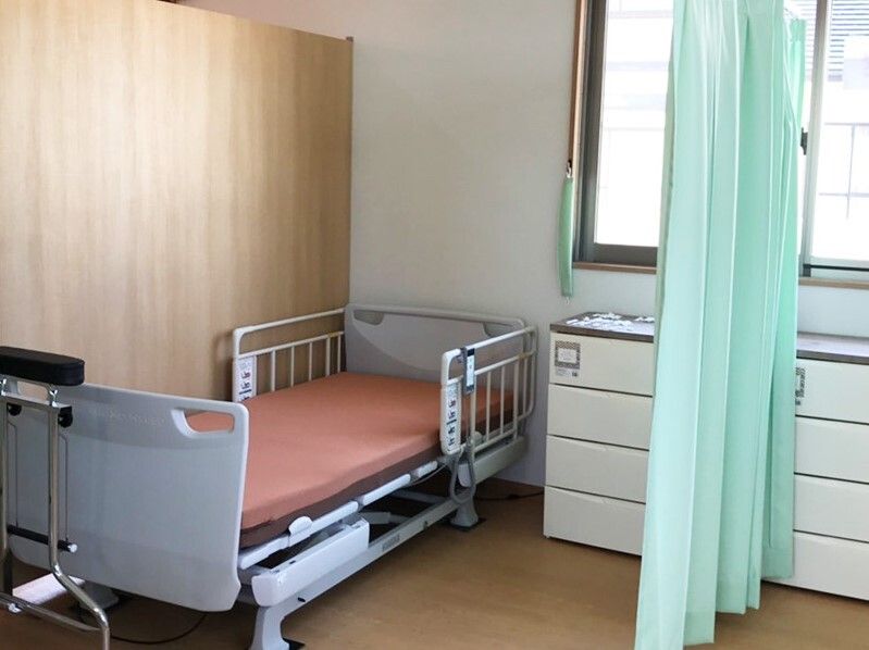 グリーンのカーテンで仕切られた部屋には介護ベッドと白いチェストが置かれていて、大きな窓も付いている。