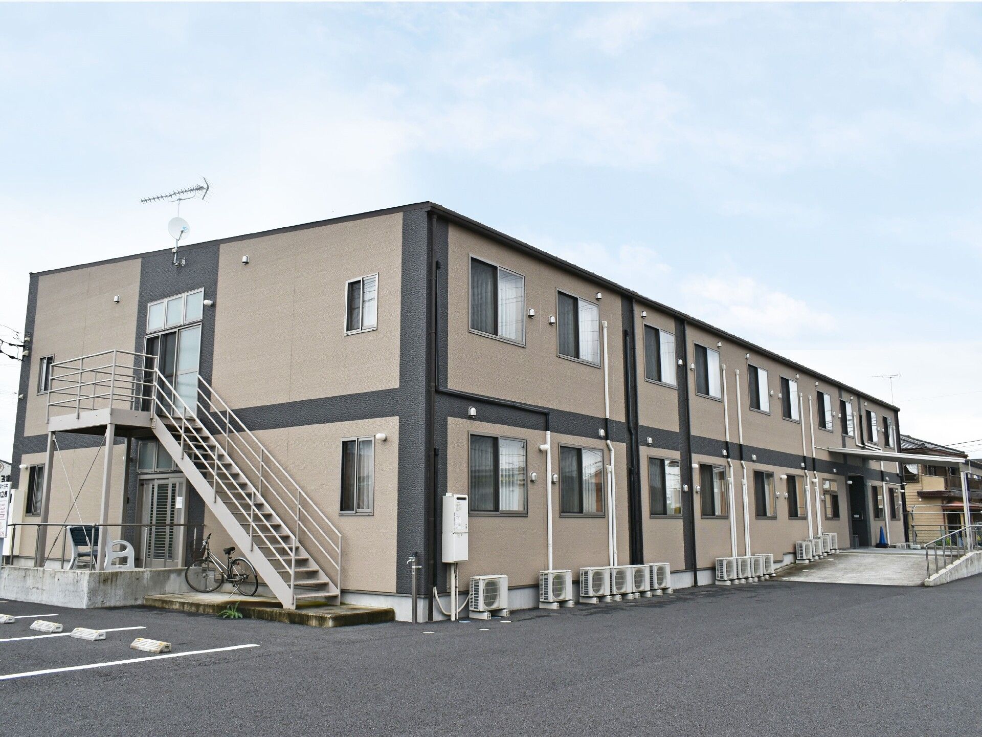 ２階建ての建物。ベージュ系で建物の前は駐車場になっている。日当たりのいい立地。