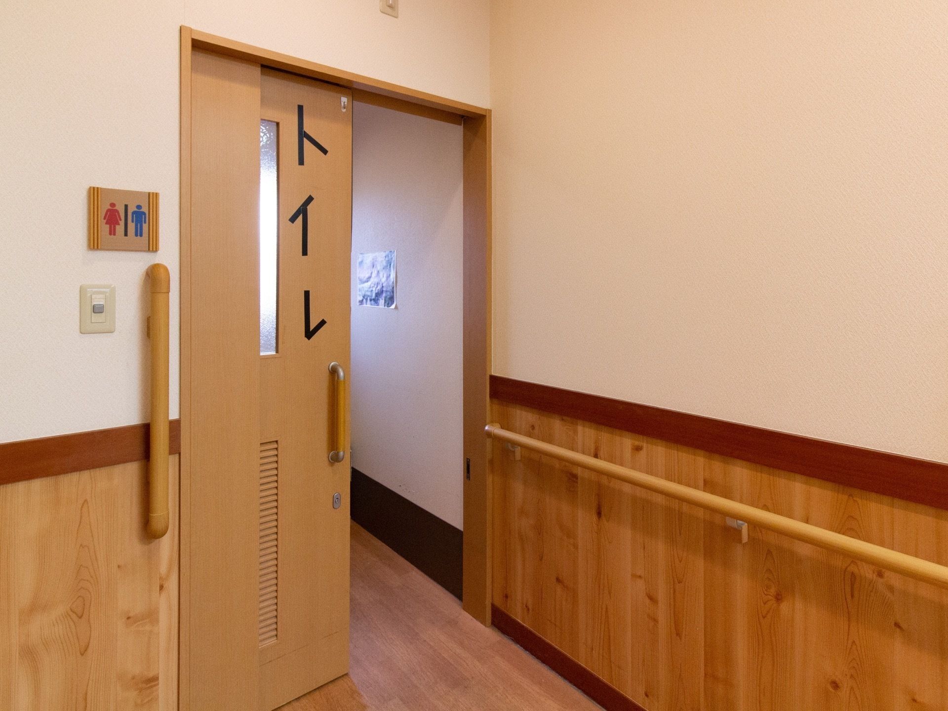 トイレの引き戸の横にはアイ字型の手すりが付き、壁にも手すりがある。「トイレ」と黒いテープで作られた文字が貼られ、壁にはトイレによくある男女のイラストも描かれている。