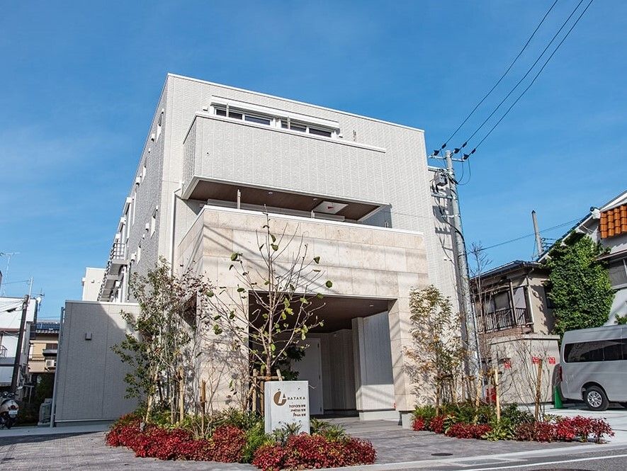 ライトグレーの3階建ての建物の2階と3階部分にはベランダがあり、周囲には住宅が建っている。建物の横には駐車場もある。