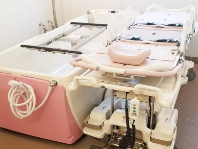 白とピンクの機械浴が設置されたスペース。右側にはストレッチャータイプのリフトがあり、左側には浴槽がある。