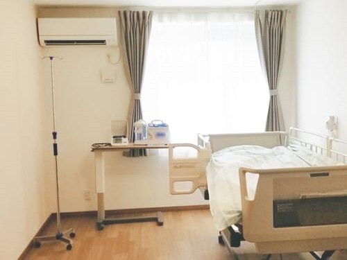 エアコンが付いたフローリングの部屋には寝具付きのベッドや緊急通報装置、カーテン付きの窓がある。