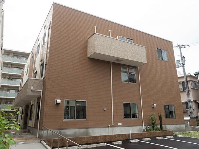 ブラウン系の３階建ての建物。玄関部分には屋根があり、手すり付きのスロープを降りると駐車場も付いている。