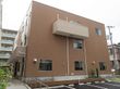 ブラウン系の３階建ての建物。玄関部分には屋根があり、手すり付きのスロープを降りると駐車場も付いている。