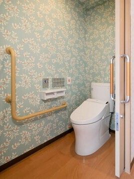 エル字型の手すりが設置されたトイレは白い花柄が描かれた空色の壁紙。オレンジの呼び出しボタンもついている。