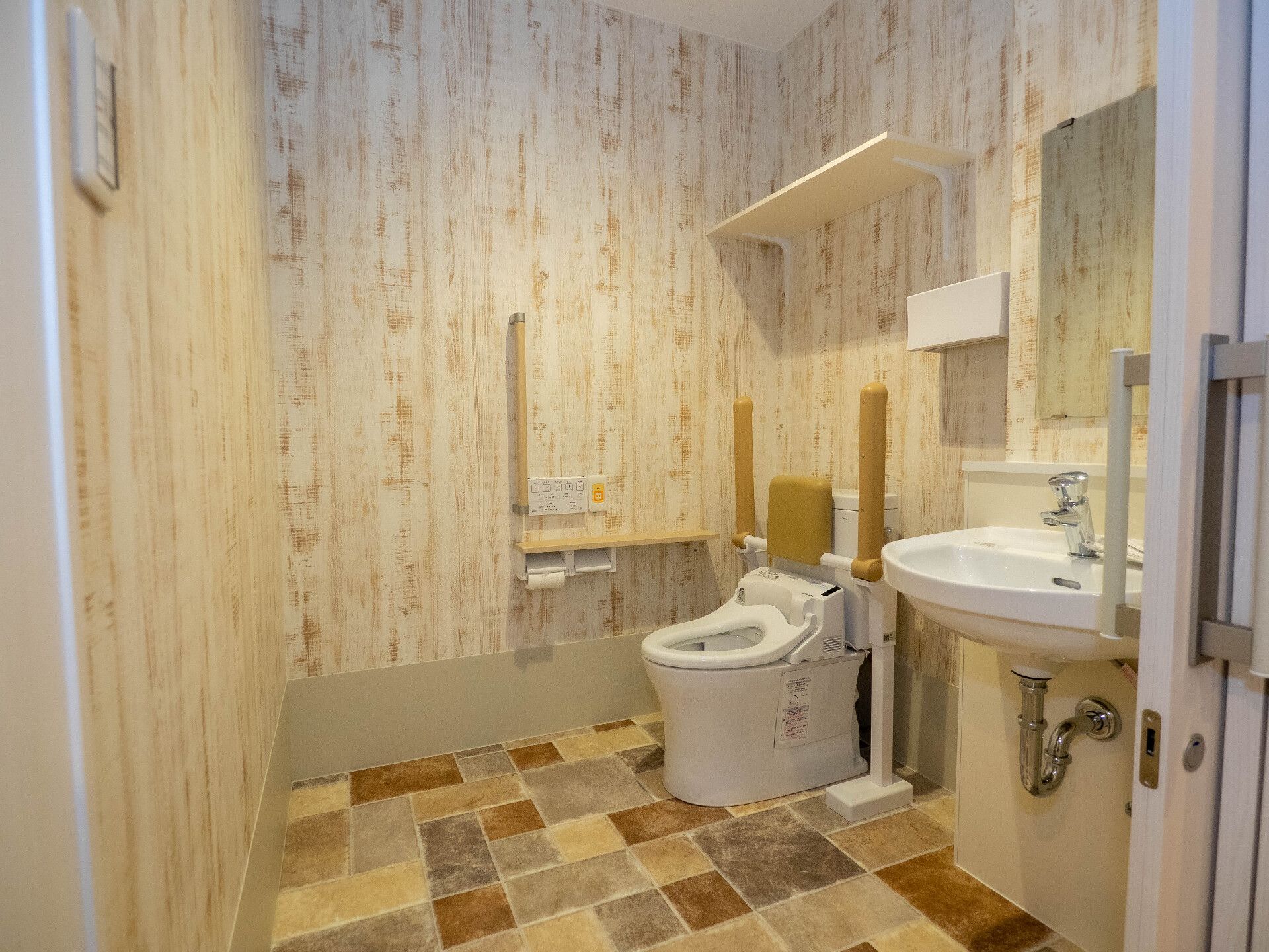 木目調の壁紙が貼られたトイレ内には手洗い場もあり、便座には背もたれと両サイドに手すりが付き、壁にもエル字型の手すりがある。