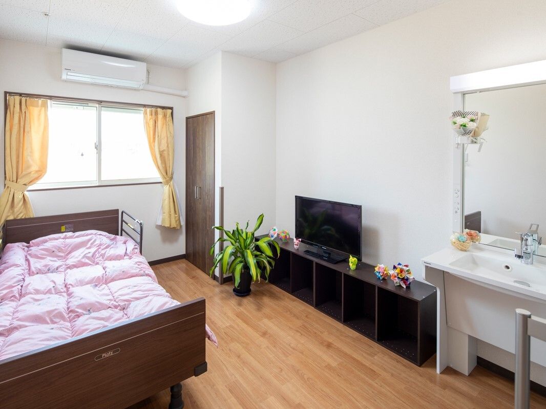 フローリングの居室にはベッドや洗面、テレビボードやテレビがあり、大きな窓にはカーテンがついている。