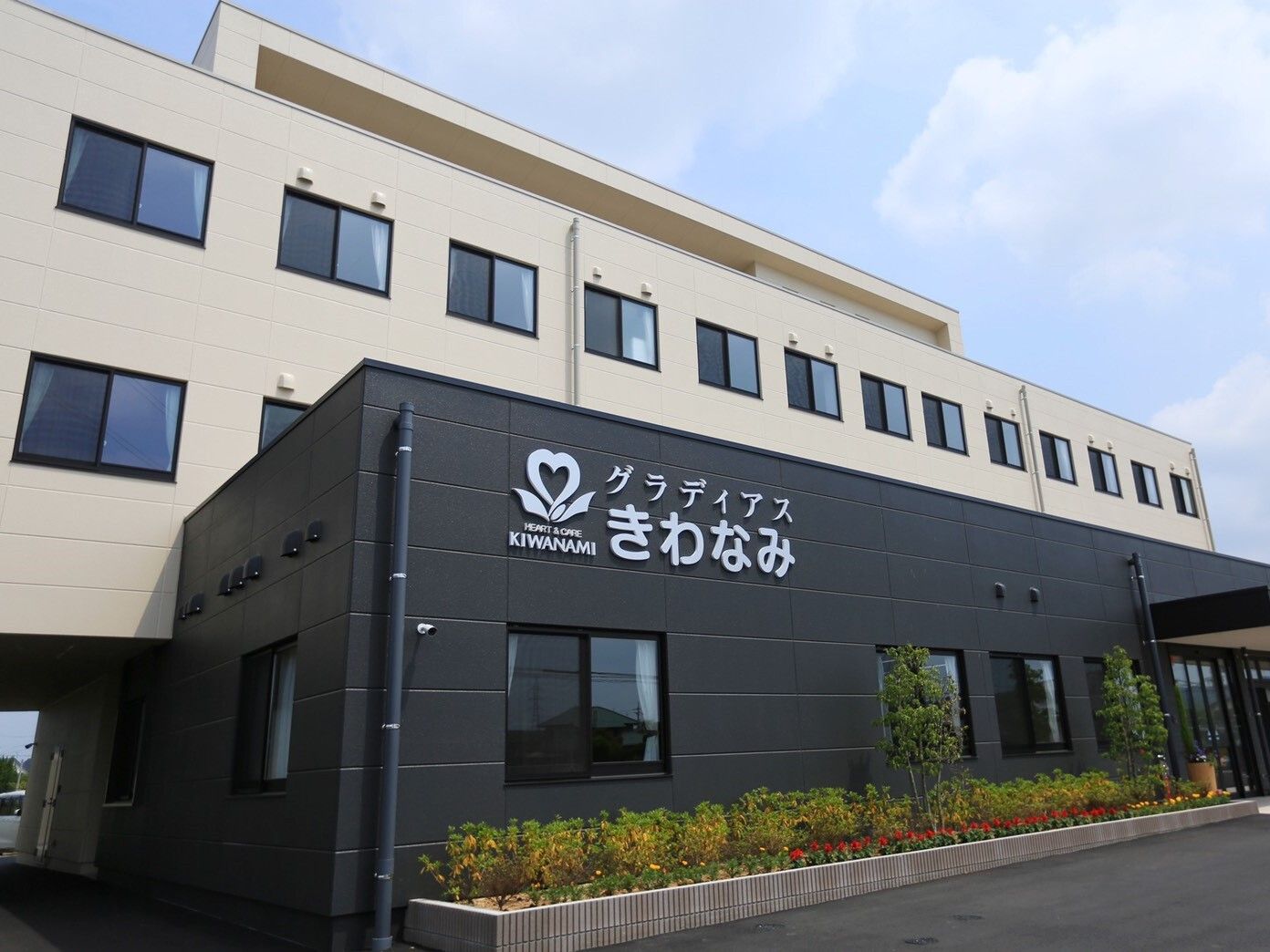 白を基調とした建物の玄関周りは黒く、すっきりとした印象。木も植えられ、外壁にはホーム名が書かれている。