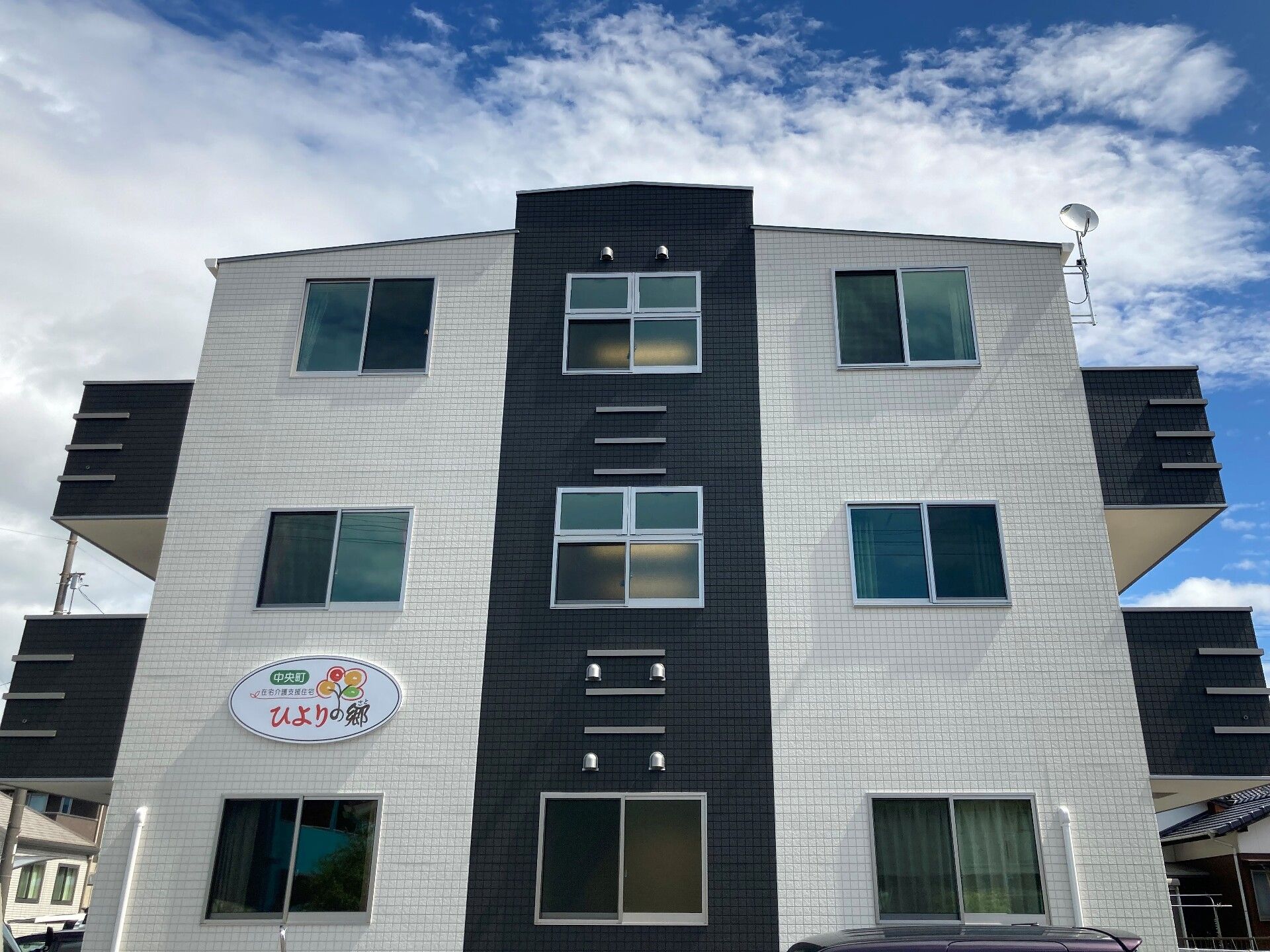 3階建ての建物は白と黒で窓がたくさんあり、各フロアにベランダも付いている。外壁には住宅名が書かれた楕円形のプレートが貼られている。