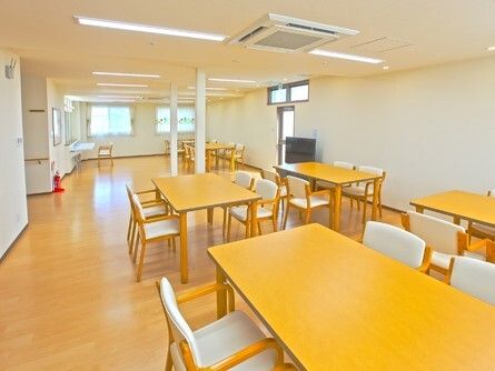 明るい色のフローリングのフロアには木製のダイニングテーブルセットが複数並び、車いす対応の洗面所や消火器、テレビなどが置かれている。