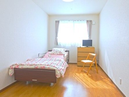 ピンクの花の寝具がセットされたベッドが置かれたフローリングの居室。テレビやチェスト、木製の椅子もあり、窓にはカーテンが付いている。