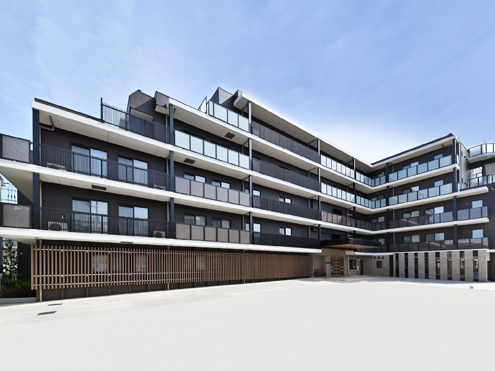 エル字型のブラウンの建物はベランダが各フロアにつき、建物の前には広い駐車場が広がっている。周辺には緑や花が植えられ、明るい雰囲気。