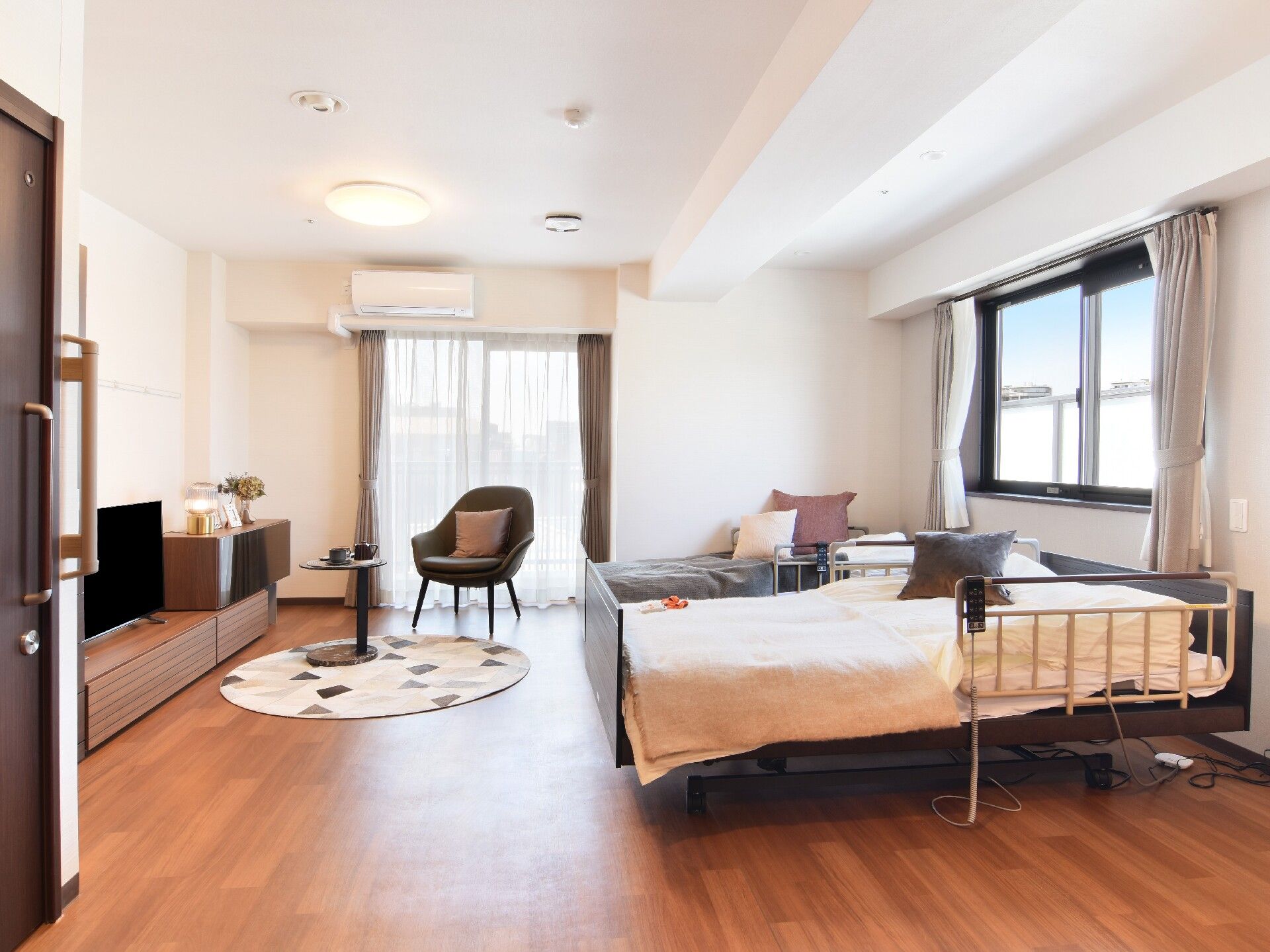 手すり付きの部屋はフローリングで、寝具付きのベッドやテレビ、テーブルセットなどが置かれ、大きな窓の外には緑が見える。