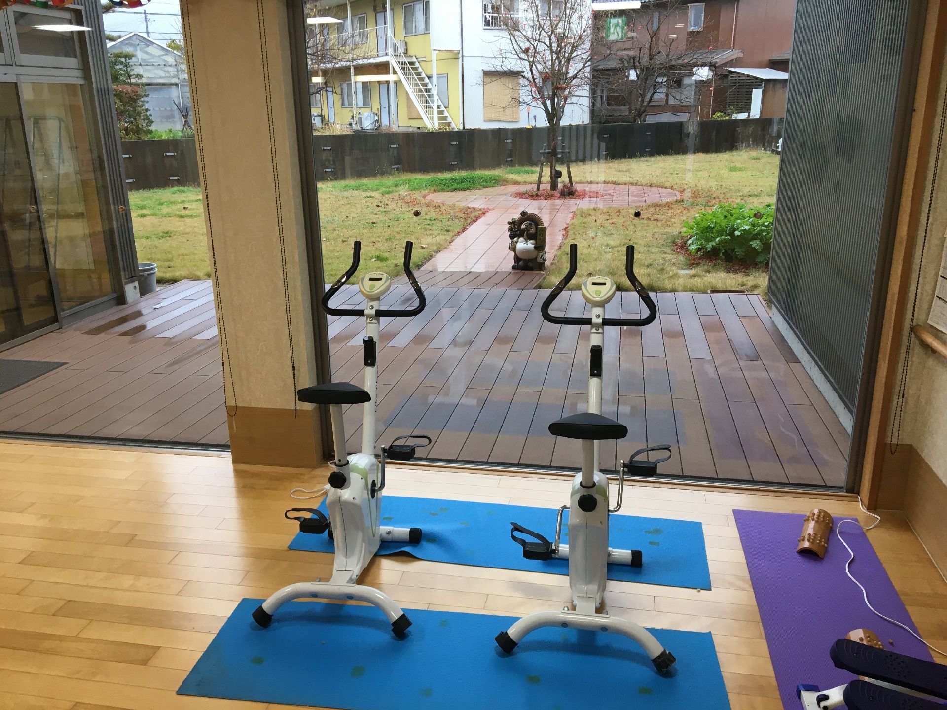 エプロンを付けた女性スタッフがニット帽を付けた車いすの高齢女性と緑の多い屋外で一緒に笑っている。