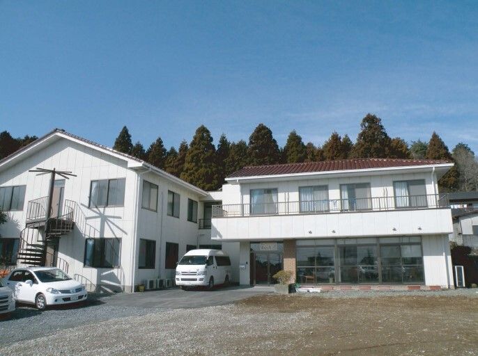 白い外壁の2階建ての建物にはエンジ色の三角屋根が付き、駐車スペースには車が止まってる。