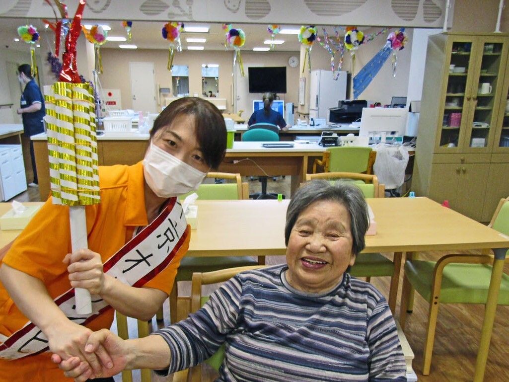 マスクをしたオレンジのポロシャツを着た若い女性スタッフが高齢女性と手を握り合っている。スタッフは「東京オリンピック」と書かれたすきをしている。