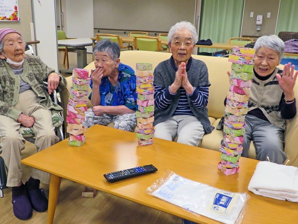 テーブルを囲んで4名の高齢女性たちがジェンガのような遊びをしている。1名は車いすに座っていて、他の3名はソファに座っている。
