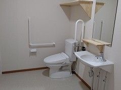 エル字型の手すりが設置された広いトイレ。鏡が付いた洗面所もあり、トイレの上には棚もある。