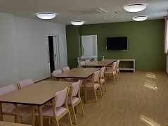 壁掛けテレビがある食堂にはピンクの椅子がセットされた木製のダイニングテーブルが複数置かれている。