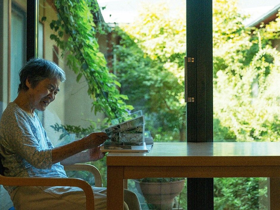 庭の見える大きな窓が付いた部屋のテーブルで雑誌を見ている高齢女性。