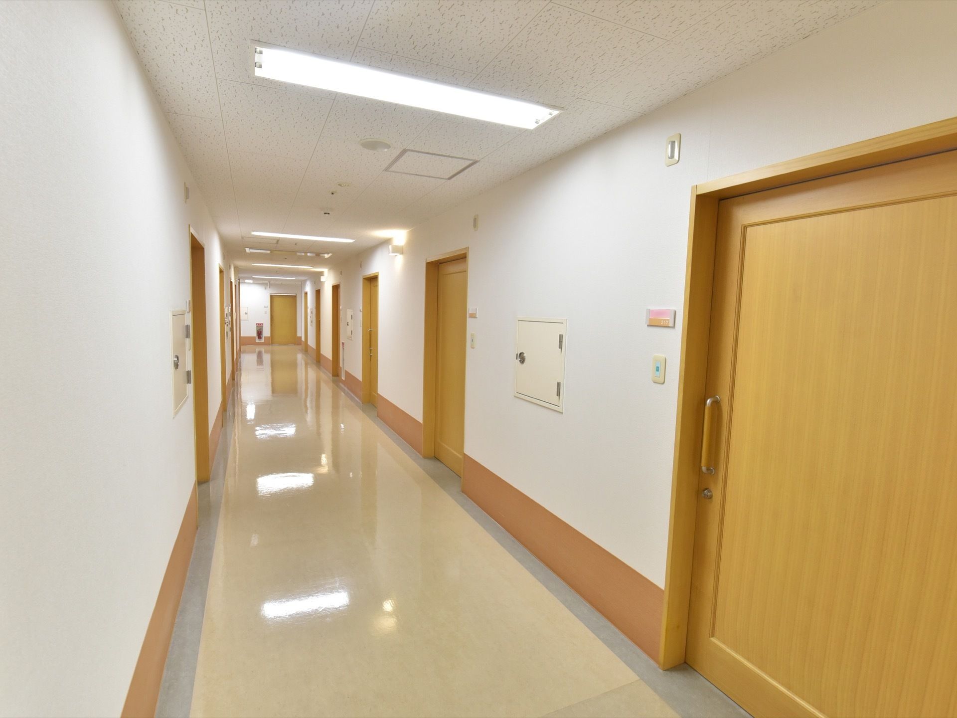 居室前の廊下である。木目調の居室扉はスライド式となっており、壁にはプライベートを考慮した呼び出しブザーが設置され、消火設備も整っている。