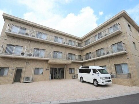 ベージュの３階建ての建物はエル字型で玄関前には白い車が止まっていて、駐車スペースもある。