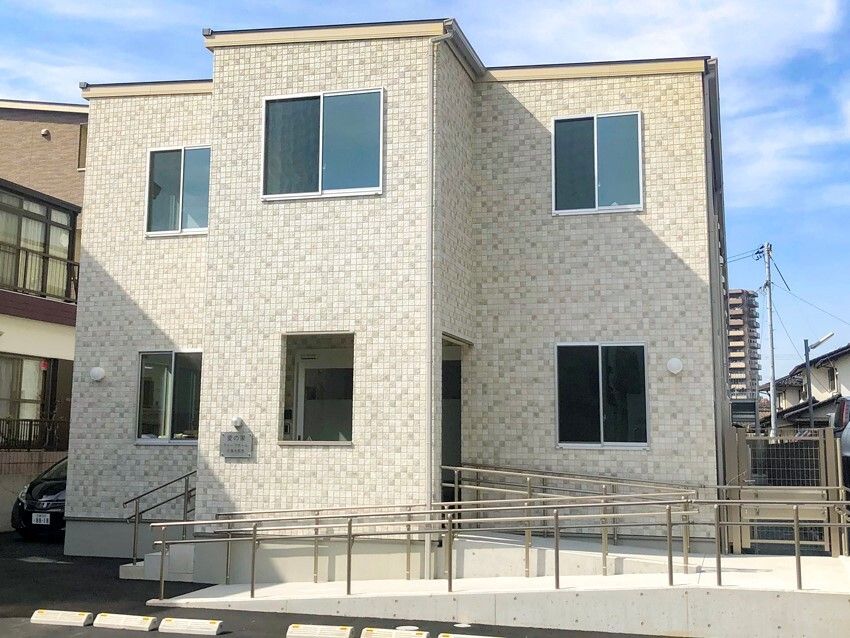 グレーがかかった白の２階建ての建物。玄関には手すりつきの階段とスロープが設置されている。
