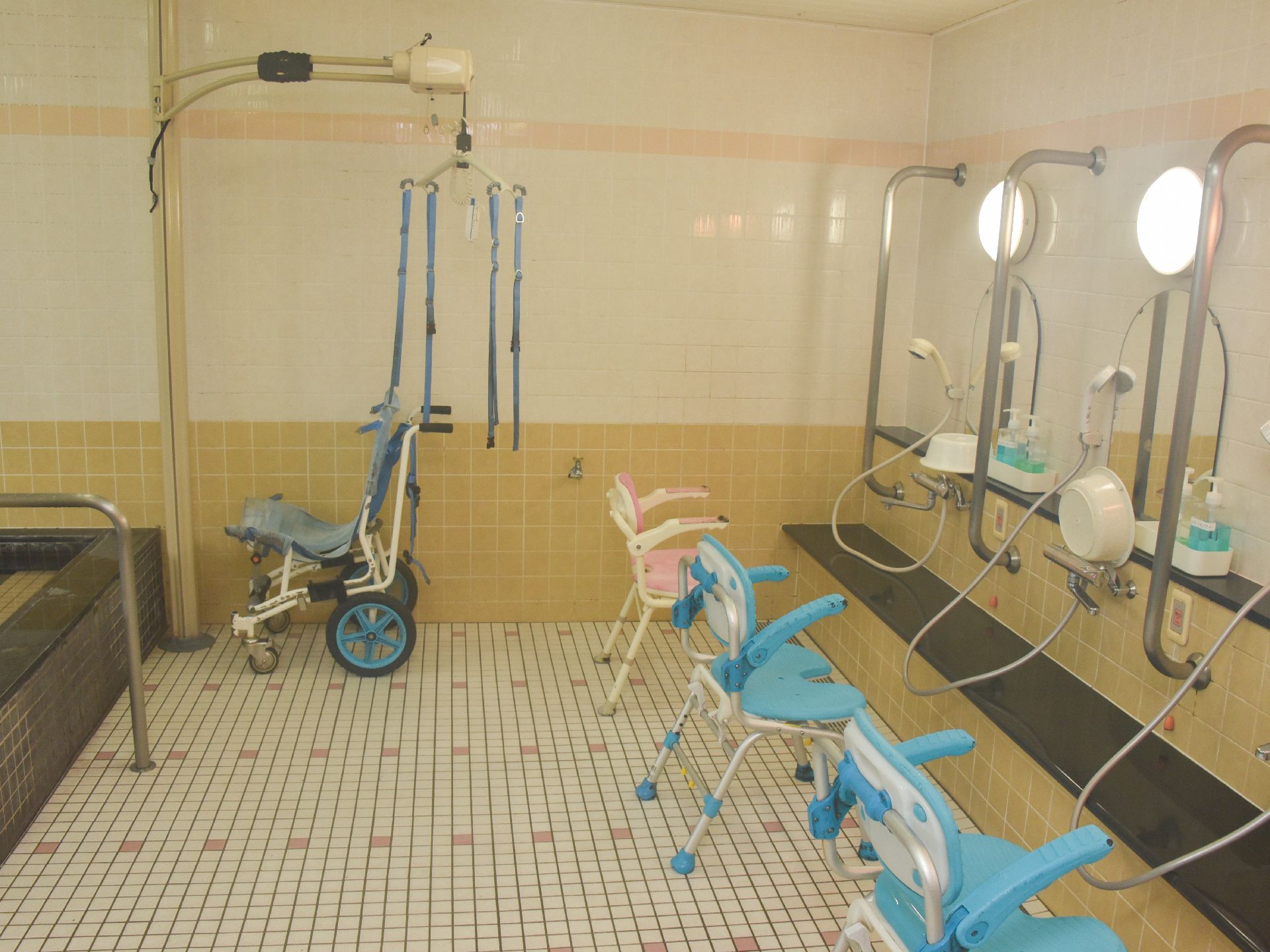 明るい浴室である。洗い場には介助用の椅子が設置されている。それぞれの鏡の左右にはステンレス製の手摺が設置されている。