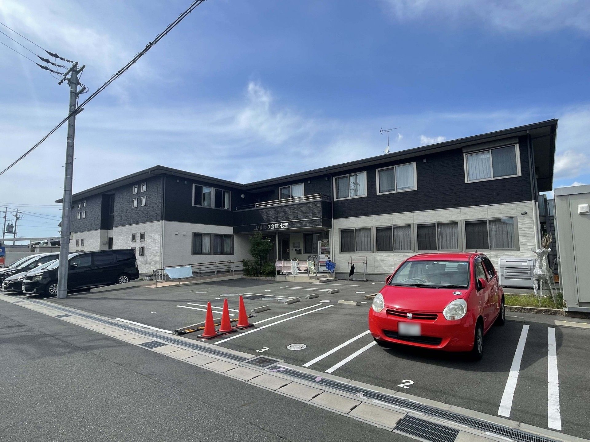 1階部分が白、2階部分がダークグレー、広い駐車場が付いた建物。玄関にはホーム名が書かれた屋根が付き、植物も植えられている。