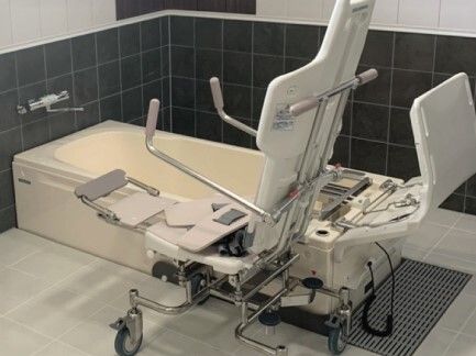 白い浴槽が設置され、３方向から介助が可能。浴槽の横には車いすのような機械が置かれ、フットレストが上がっている。