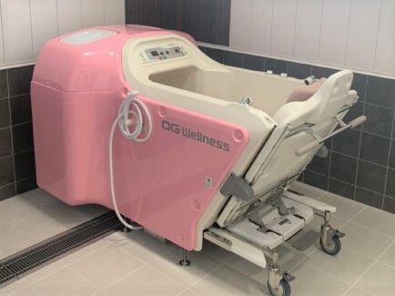 座ったまま入浴できる白とピンクの機械浴が置かれたタイル張りの浴室。シャワーが側面に設置されている。
