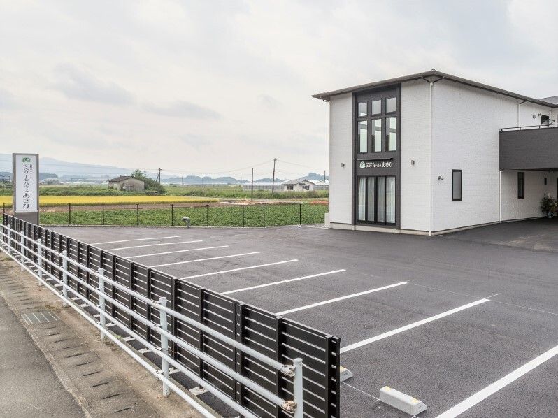 白と黒の2階建ての建物が立つ敷地内には白線が引かれた駐車場があり、周りには田畑が広がっている。