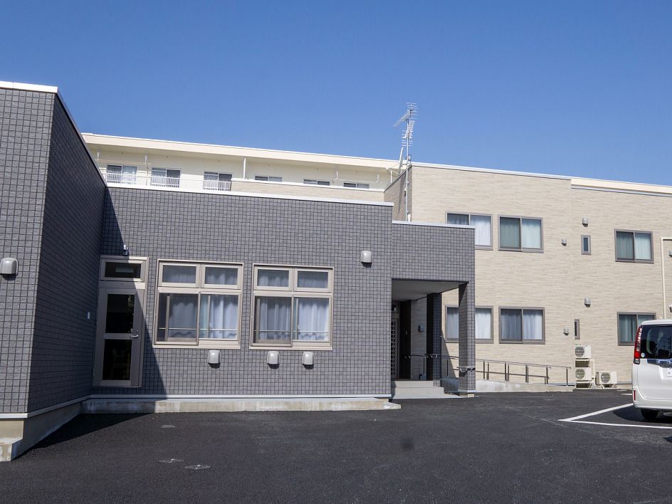 白を基調とした2階建ての建物は一部平屋建てで、周辺には木が植えられ、白いフェンスもある。