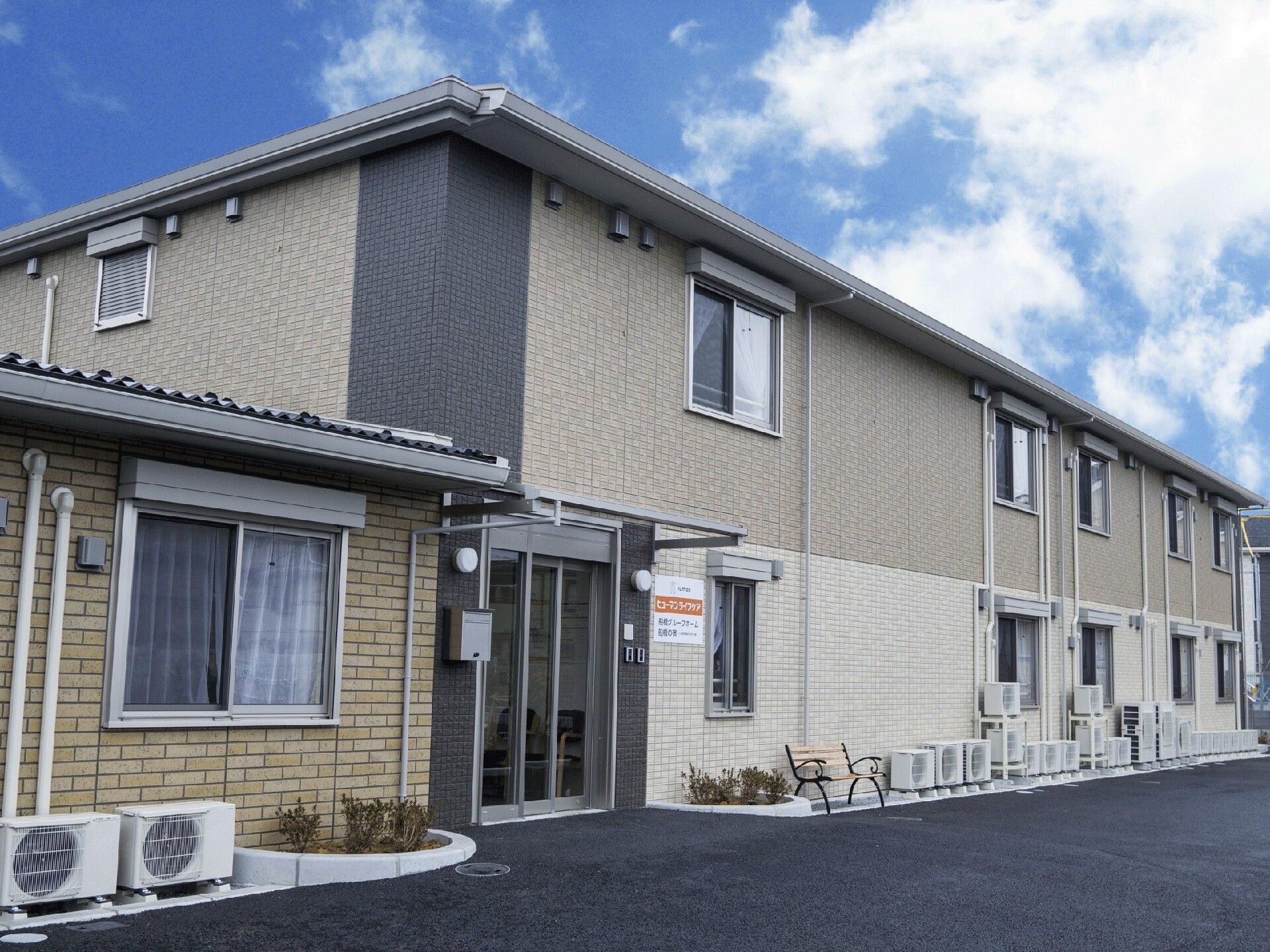 広い駐車場があり、たくさんの窓が付いた建物。ブラウン系の２階建ての建物は手前の一部建物は平屋建てで、黒っぽい屋根が付き、明るい雰囲気。