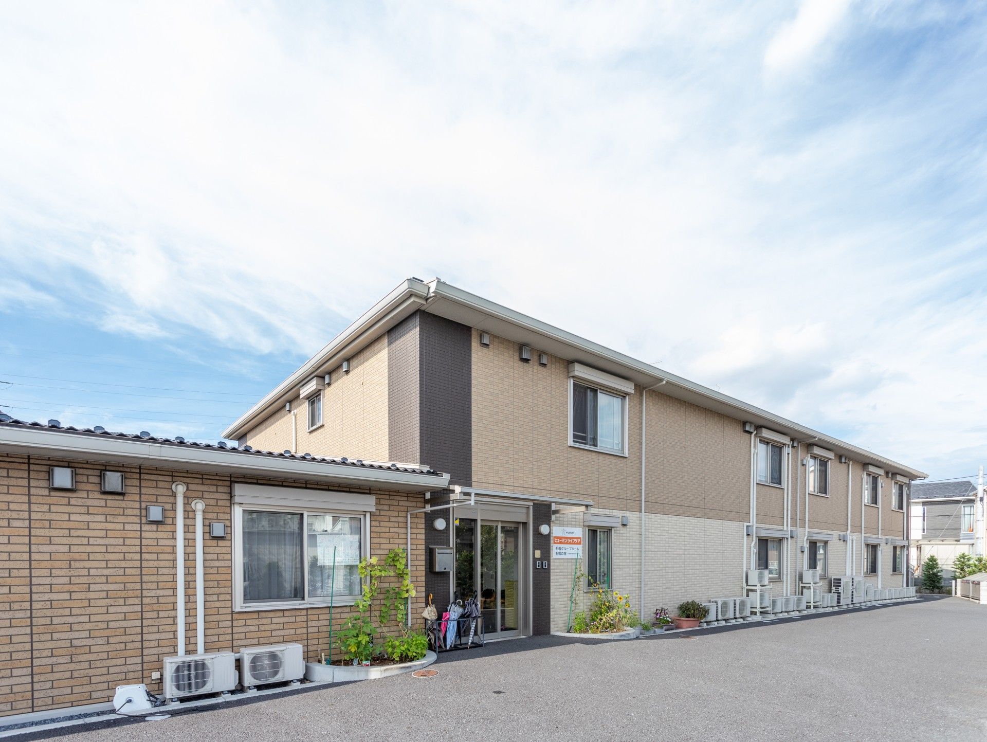ブラウン系の建物は手前が平屋建て、奥が2階建てで、駐車場も付いている。敷地の周りには白いフェンスもある。